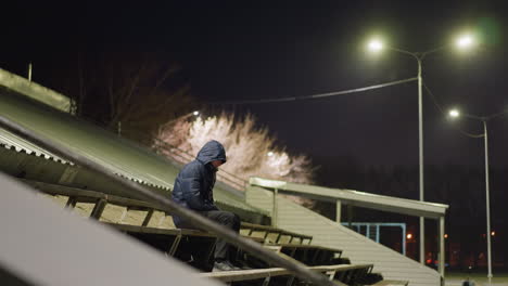 a man wearing a hooded jacket sits alone on stadium bleachers at night, illuminated by streetlights, the background includes a dimly lit building, flying particles, and a distant passing car