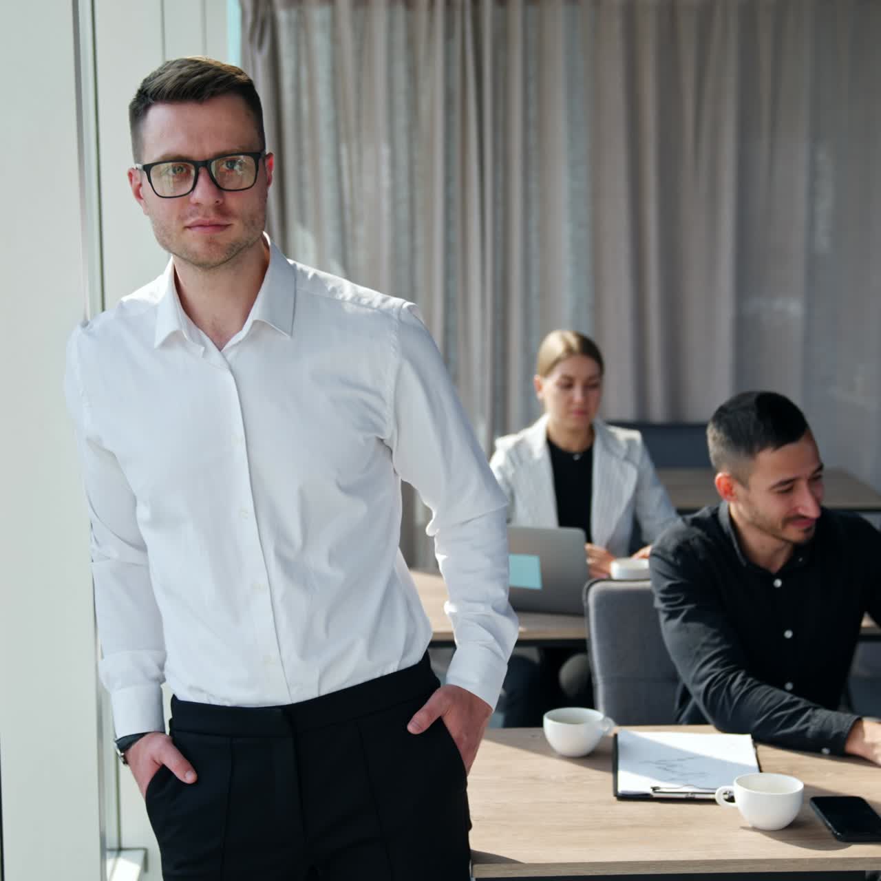 Calm peaceful pensive man standing at the window thinking over something. Colleagues working at laptops at backdrop