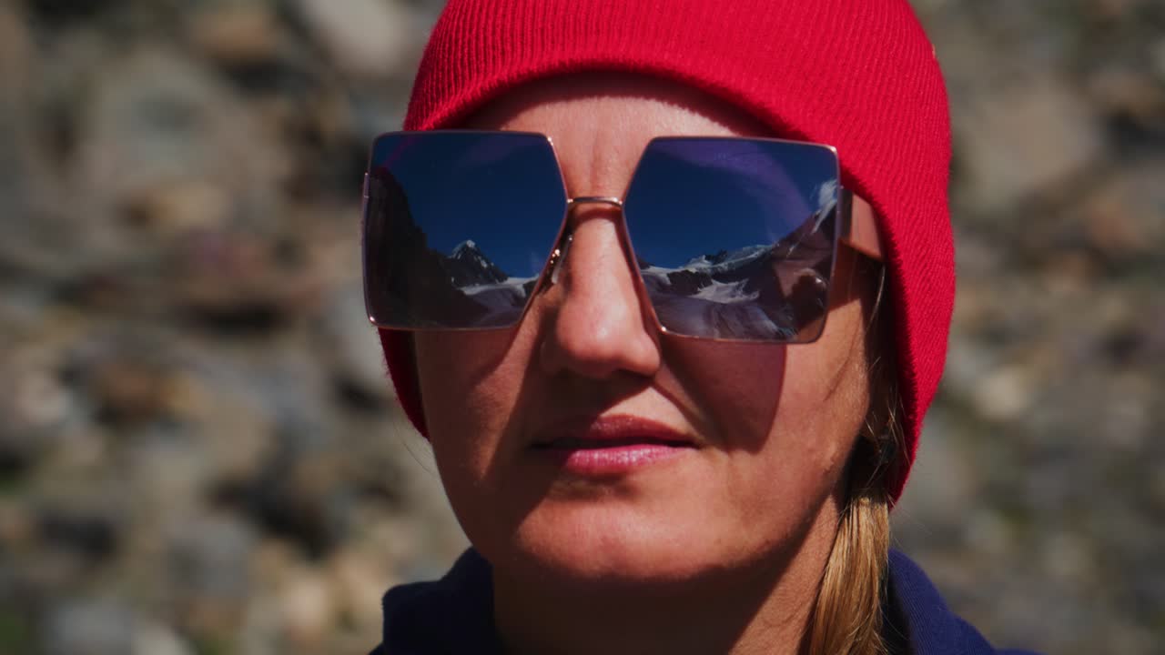 Woman in red beanie and sunglasses stands against rocky mountain backdrop. She gazes calmly, reflecting snowy peaks in her lenses. Outdoor adventure and exploration theme suits travel, fashion