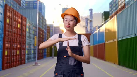 trabajadora asiática con gafas de protección y casco de seguridad mirando a la cámara y mostrando el gesto de las manos mientras está de pie en el almacén del patio de contenedores