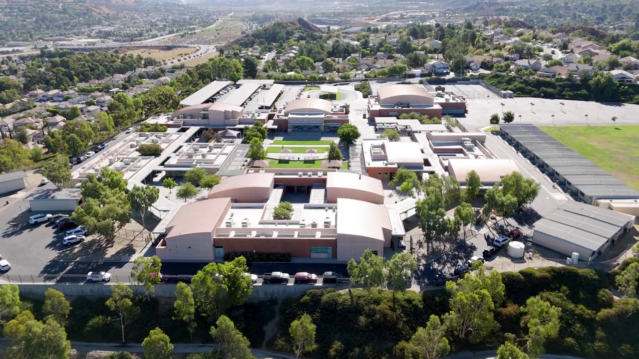 Edifícios de escola secundária de Santa Clarita, campos esportivos, layout moderno, vegetação abundante e paisagem suburbana sob a luz do sol - revelação aérea ascendente.