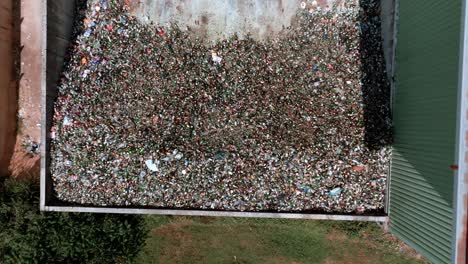 Vista-Aérea-Ascendente-De-Drones-Desde-El-Centro-De-Reciclaje-De-Vidrio-Del-Almacén,-Siguiendo-A-Una-Excavadora-De-Montacargas-Agarrando-Vidrio-Y-Cargándolo-En-La-Parte-Trasera-De-Un-Camión-Transportador,-Concepto-De-Reciclaje-De-Vidrio