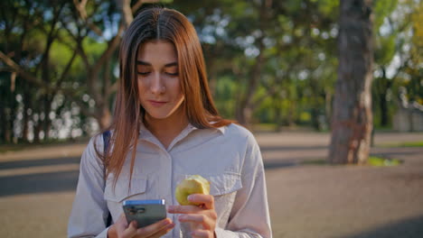 mujer comprometida mirando teléfono inteligente sosteniendo manzana en verde parque soleado primer plano.