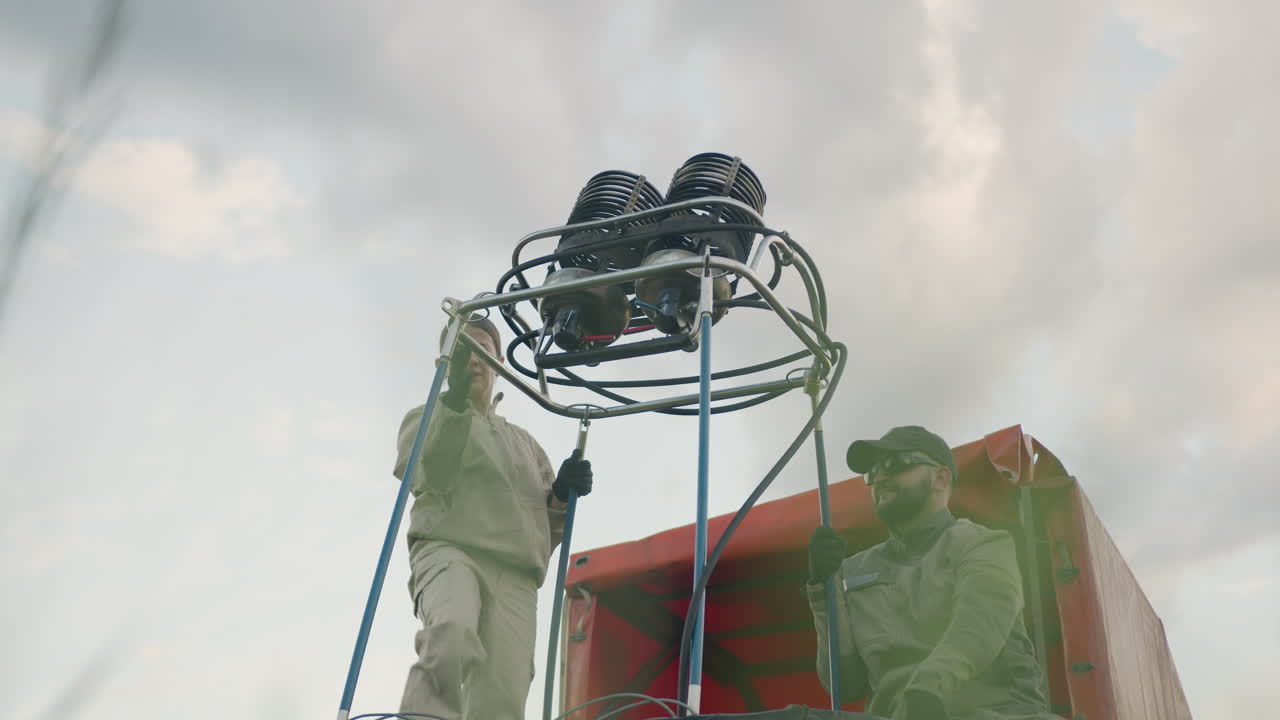 Woman in beige jumpsuit stands inside basket fixing metal burner unit while man assists by steadying vertical poles under cloudy sky during balloon assembly process in outdoor open field