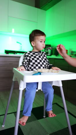 beautiful dark-haired baby boy sits at high chair in the home kitchen. kid looks focused ahead while mom feeds him from spoon. vertical video.