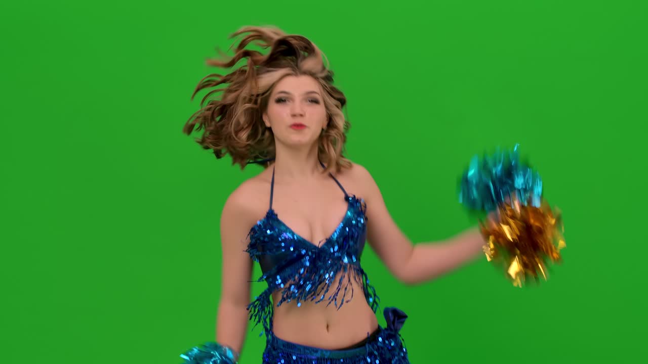 retrato de una joven animadora feliz bailando en el estudio en una pantalla verde. una chica en un traje de baño azul realiza elementos con pom-poms, entrenando antes de la competencia. cámara lenta. primer plano