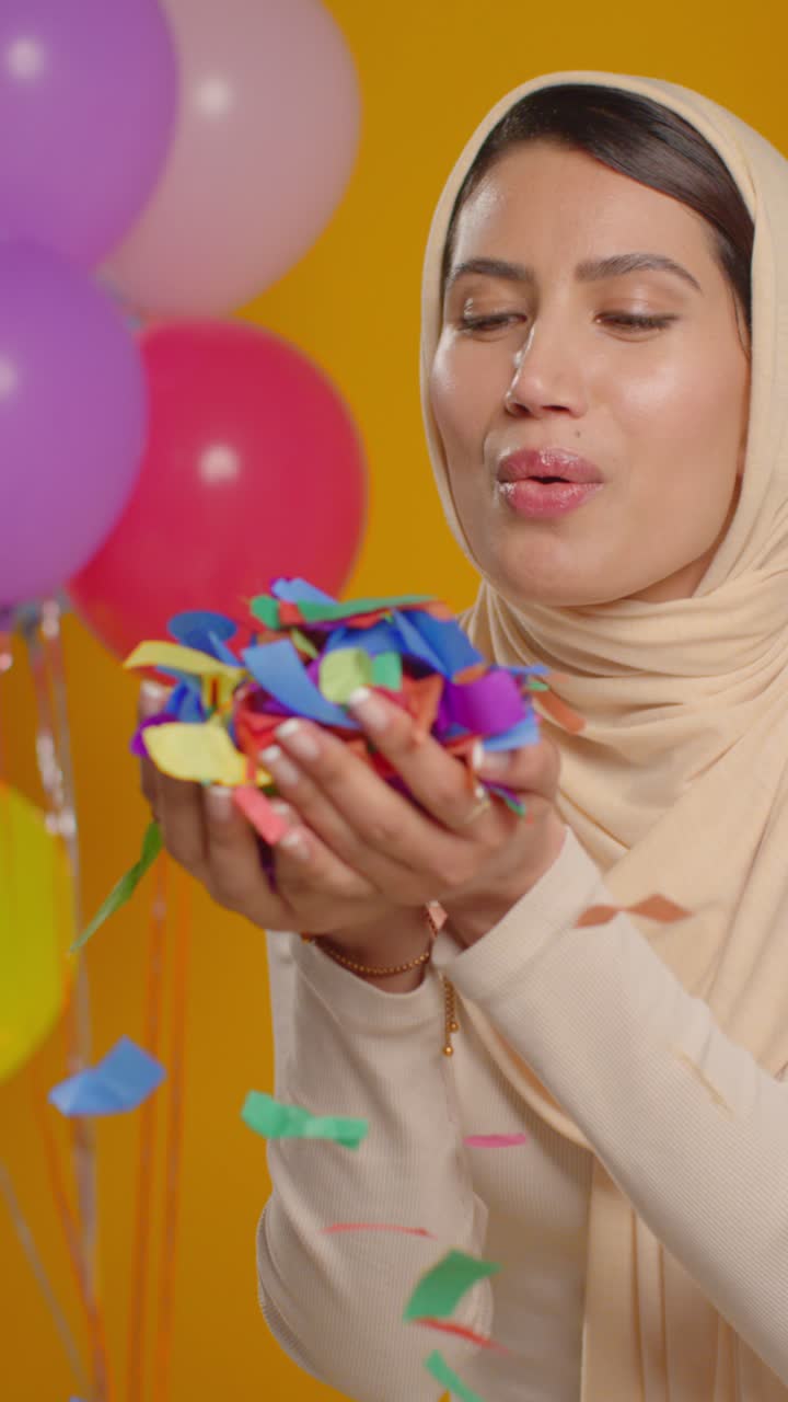 retrato vertical de estudio de video de una mujer con hijab celebrando su cumpleaños soplando confeti de papel rodeado de globos 1