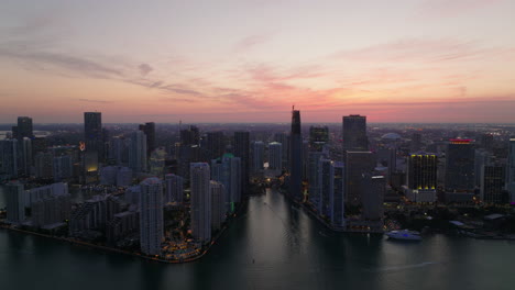 Backwards-reveal-of-modern-high-rise-downtown-buildings-on-waterfront-at-twilight.-Beautiful-view-of-urban-borough-under-pink-sky.-Miami,-USA