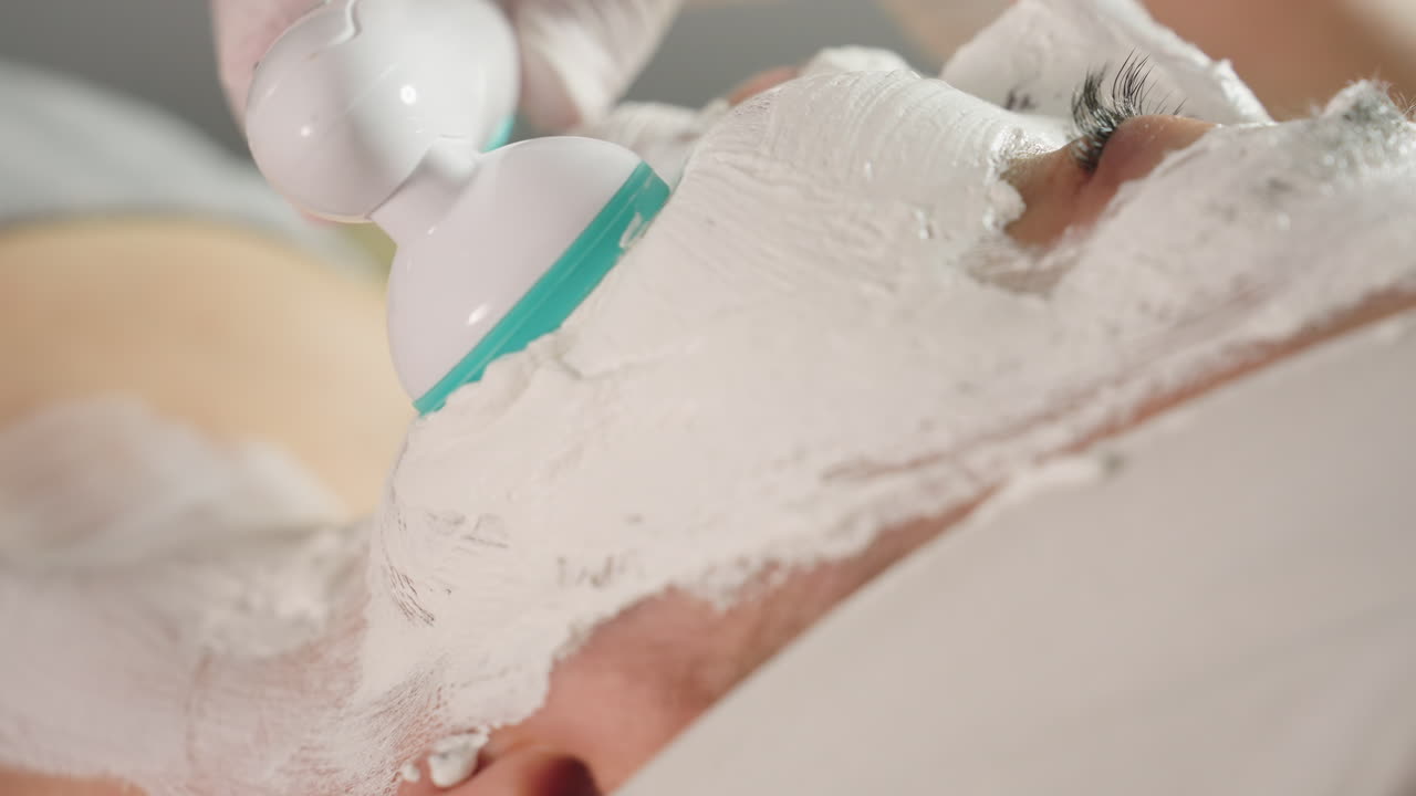 Skin care specialist uses vibrating electric massager to smooth thick white cream mask on spa guest face and jawline in close up for deep rejuvenation and soothing relaxation under soft clinic light