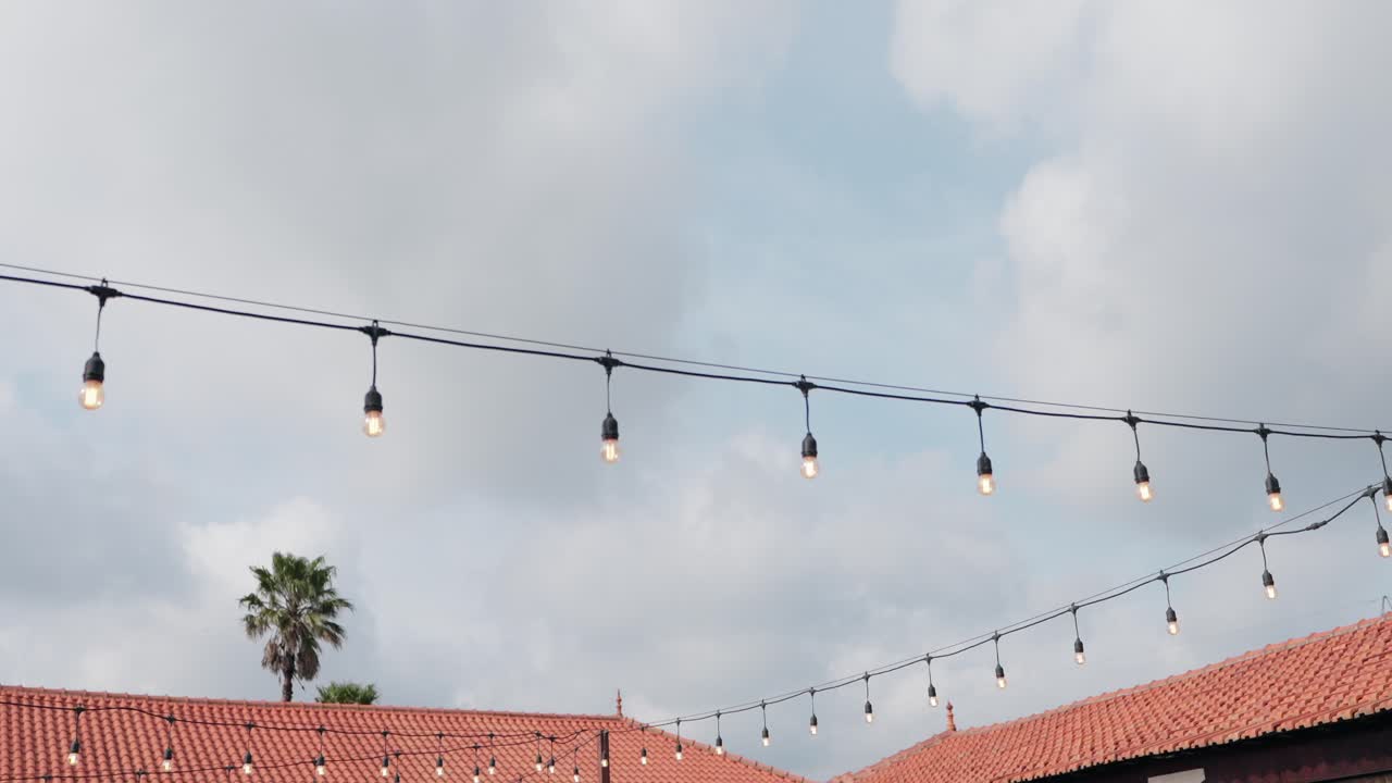 Outdoor string lights hanging above red tiled rooftops with a cloudy sky and palm tree
