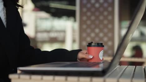vista lateral de la computadora portátil en la mesa con un teclado visible, persona que sostiene una taza de café cerca de la laptop, la deja caer en la mesa y comienza a escribir, adornada con anillos, entorno de restaurante moderno con fondo borroso