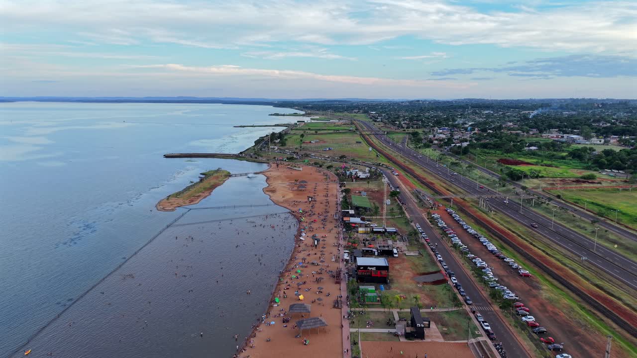 Ripresa panoramica a ritroso con drone sopra la popolare spiaggia locale Balneario Costa Sur, con strada statale che corre parallela al tranquillo fiume Paraná, Posadas, Misiones, Argentina