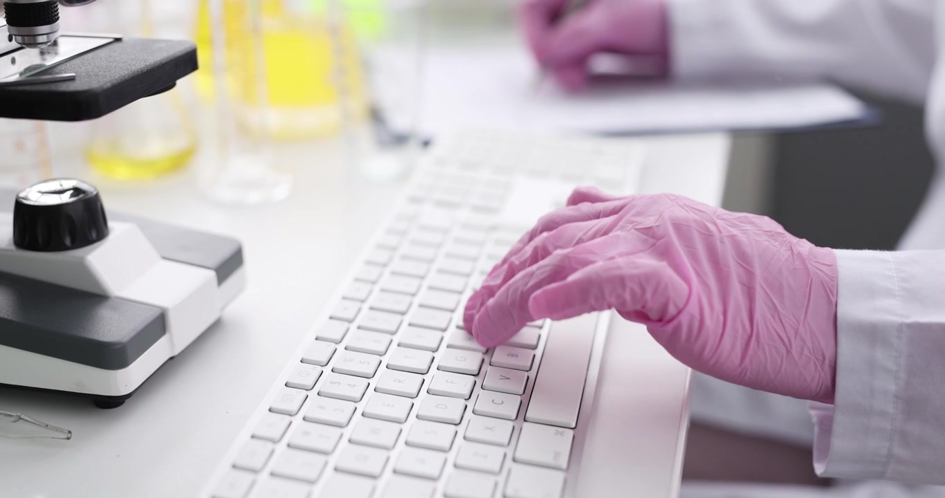 Person wearing gloves inputs lab data on keyboard near microscope
