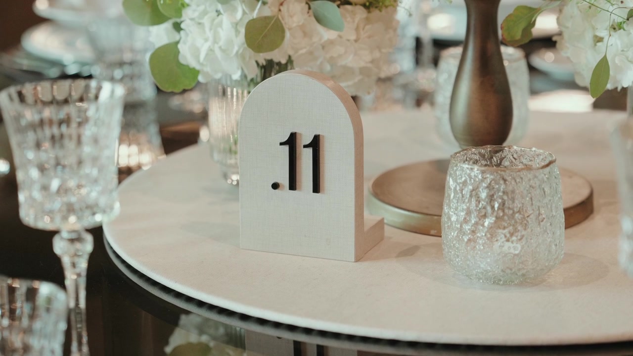 Letrero de número de mesa arqueado sobre una mesa con espejo junto a copas de cristal y flores blancas