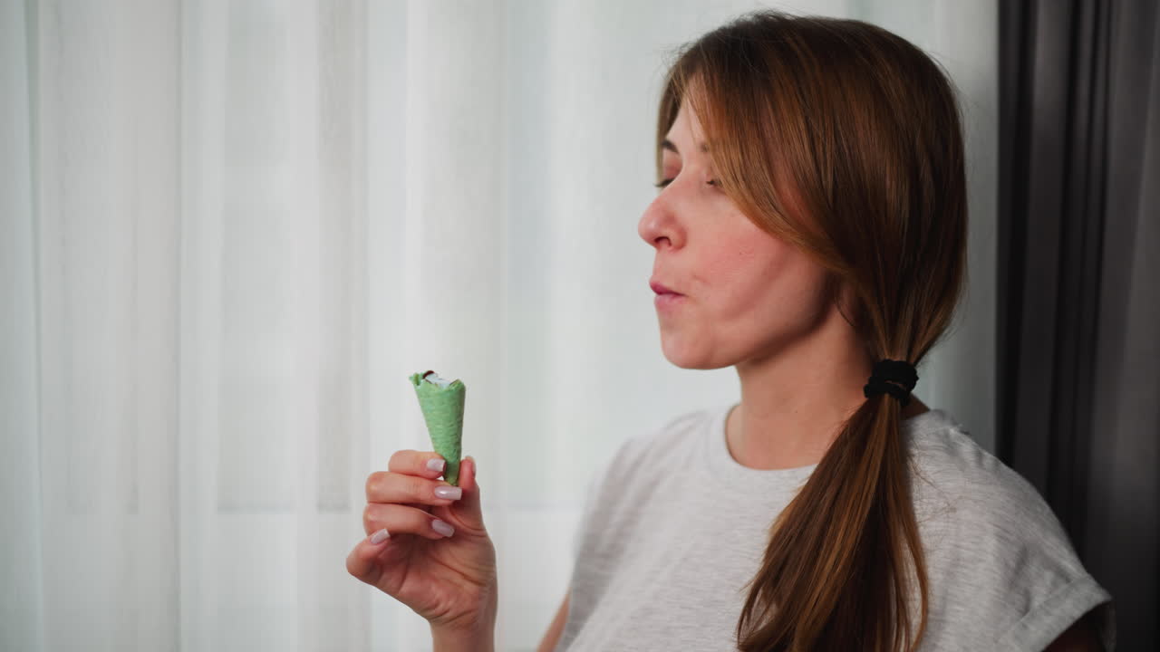 Portrait view of calm woman with ponytail hair wearing grey top savoring delicious ice cream from green cone standing peacefully by sheer curtain in cozy bright