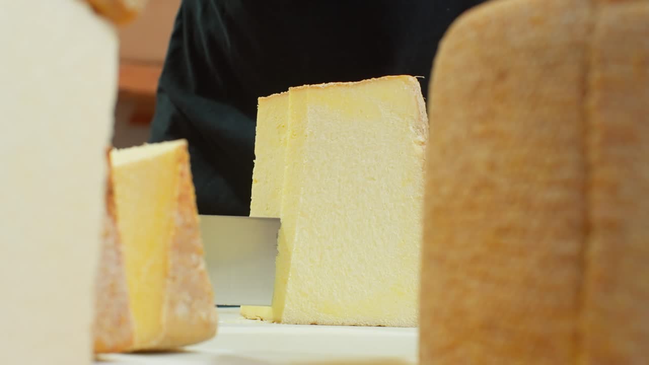 A large cheese block is sliced smoothly with a sharp tool in a dairy setting