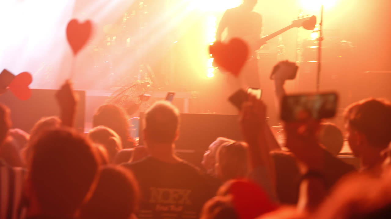 Red heart shaped signs waving, audience members recording live music concert moment with smartphones, capturing shared passion and excitement of musical performance