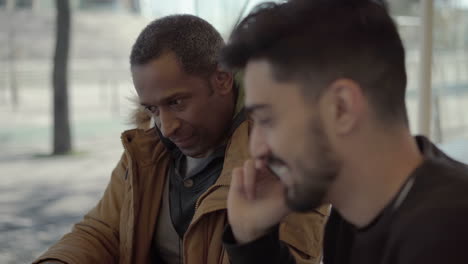 smiling man talking by smartphone and sitting with friend in cafe