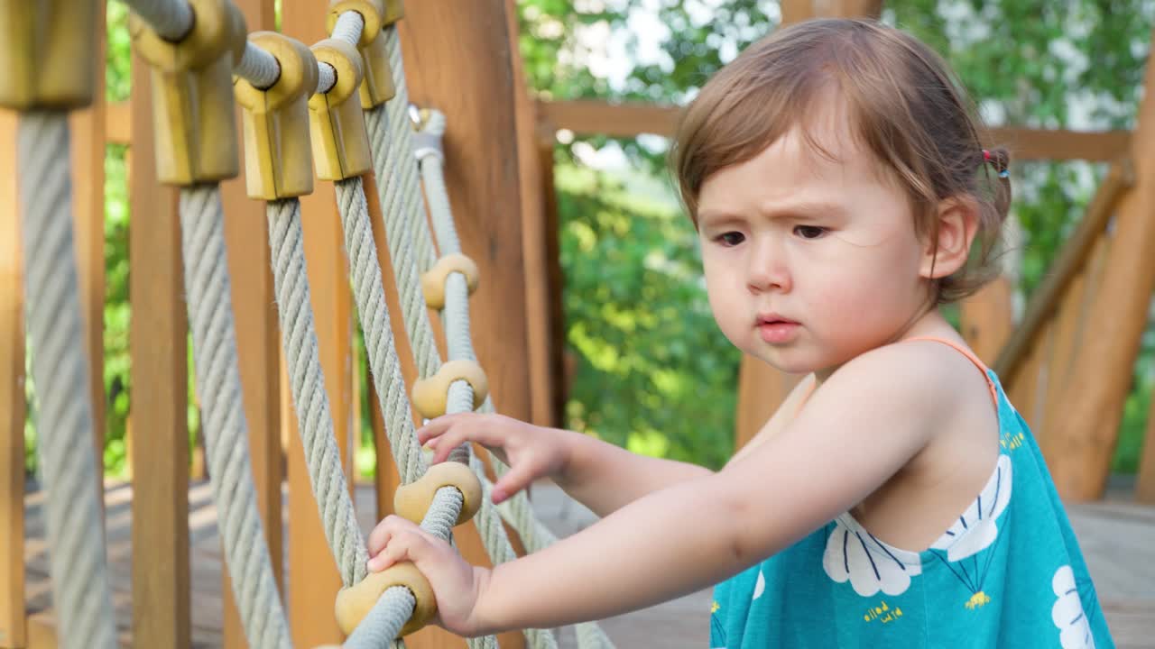 Adorável menina atravessando uma ponte de madeira segurando cordas com as duas mãos em um playground ao ar livre - vista lateral em câmera lenta.
