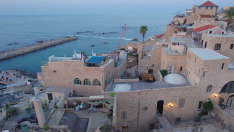 Los-Hermosos-Y-Antiguos-Edificios-De-La-Antigua-Ciudad-De-Jaffa-Al-Atardecer-Con-Gente-Sentada-En-Los-Tejados-Para-Mirar-La-Puesta-De-Sol-Con-Los-Barcos-Del-Puerto-De-Jaffa-Al-Costado-Del-Marco---Disparo-De-Drones-De-Paralaje