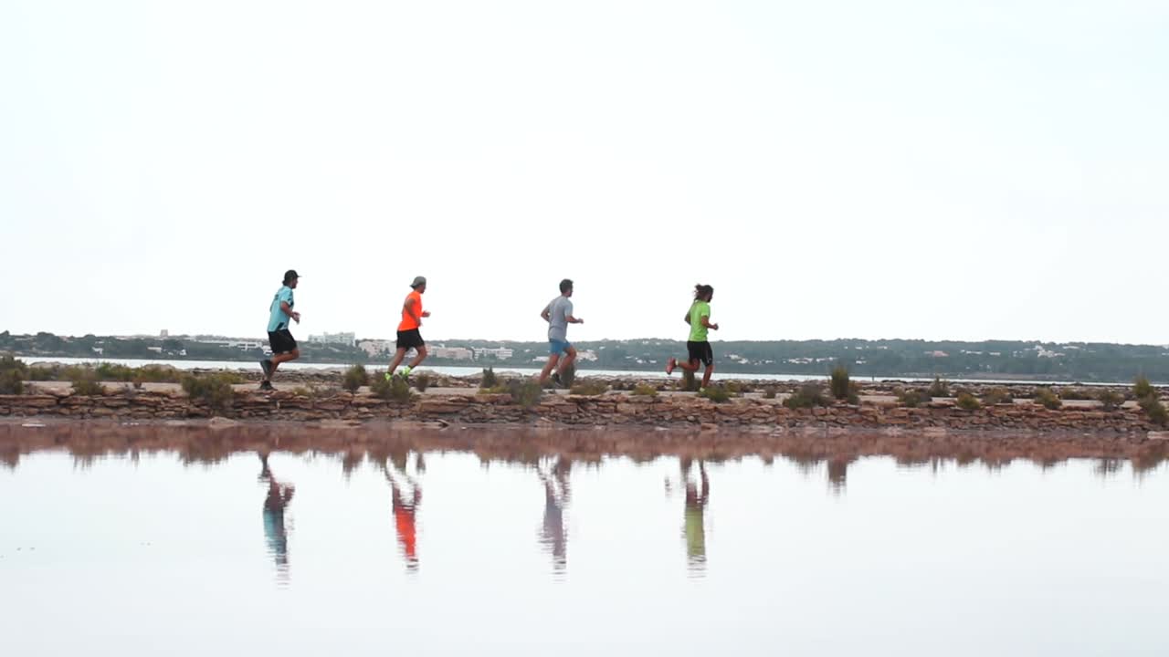 Smooth pan shot of four men running behind each other in front of a lake in Formentera. You can see its reflections in the water, and behind some beautiful houses