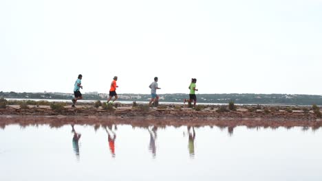 smooth pan shot of four men running behind each other in front of a lake in formentera. you can see its reflections in the water, and behind some beautiful houses