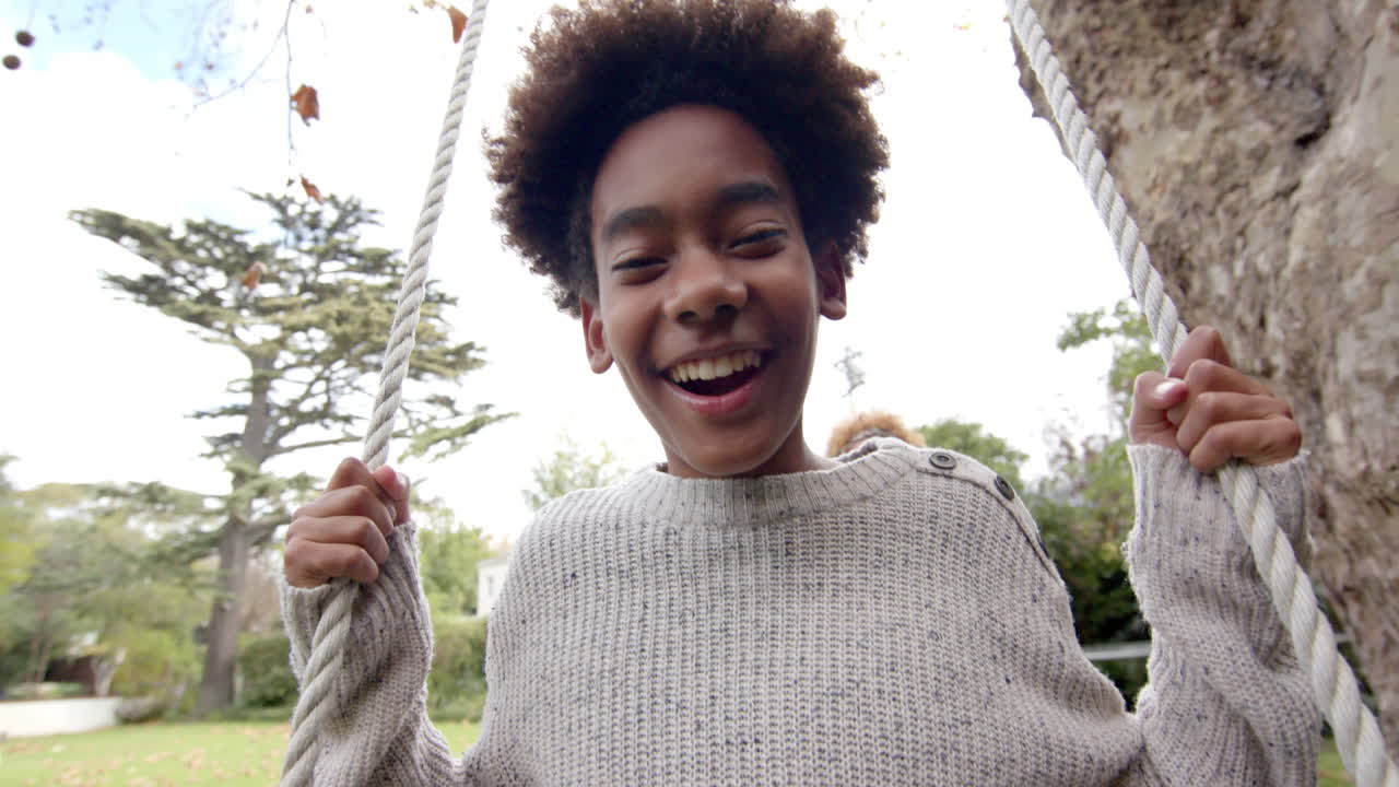 Portrait of happy african american son on tree swing with mother pushing, slow motion