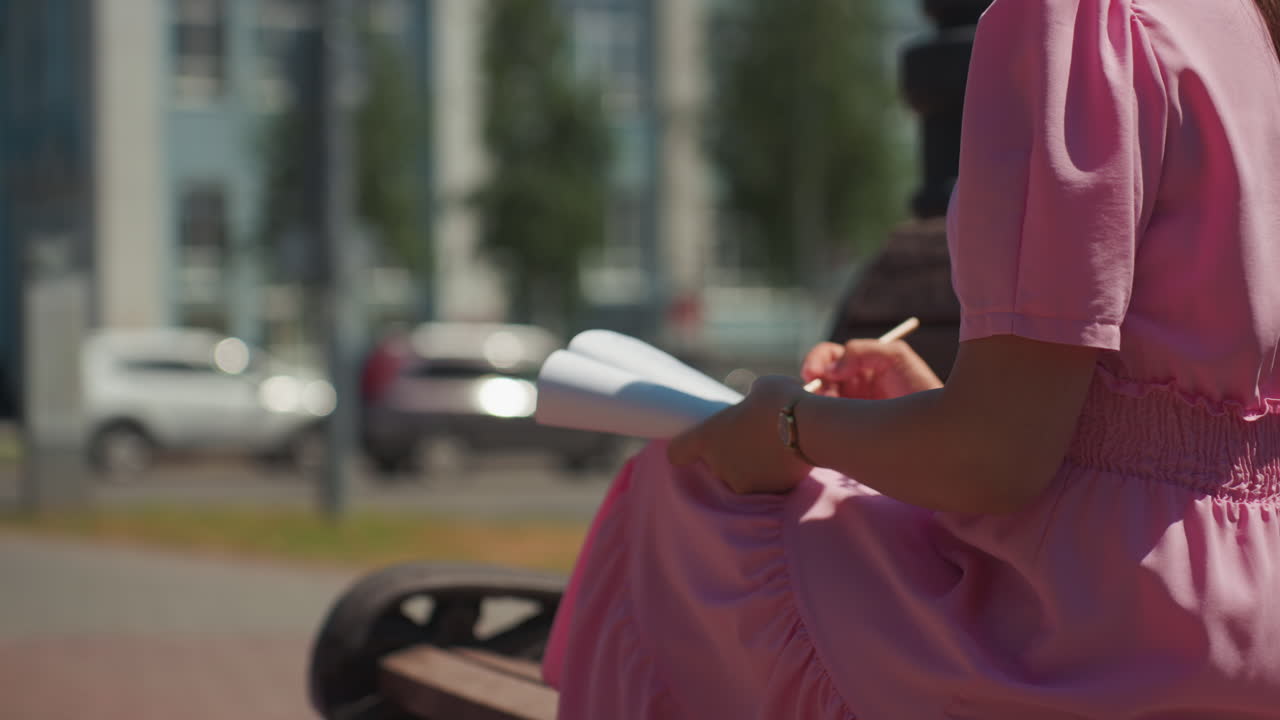 Kvinde der tager noter på en bænk, beskåret billede af siddende skribent der noterer idéer i notesbog med blyant, byens gade og træer sløret i baggrunden, pink kjole og afslappet fokuseret kropsholdning, naturligt dagslys