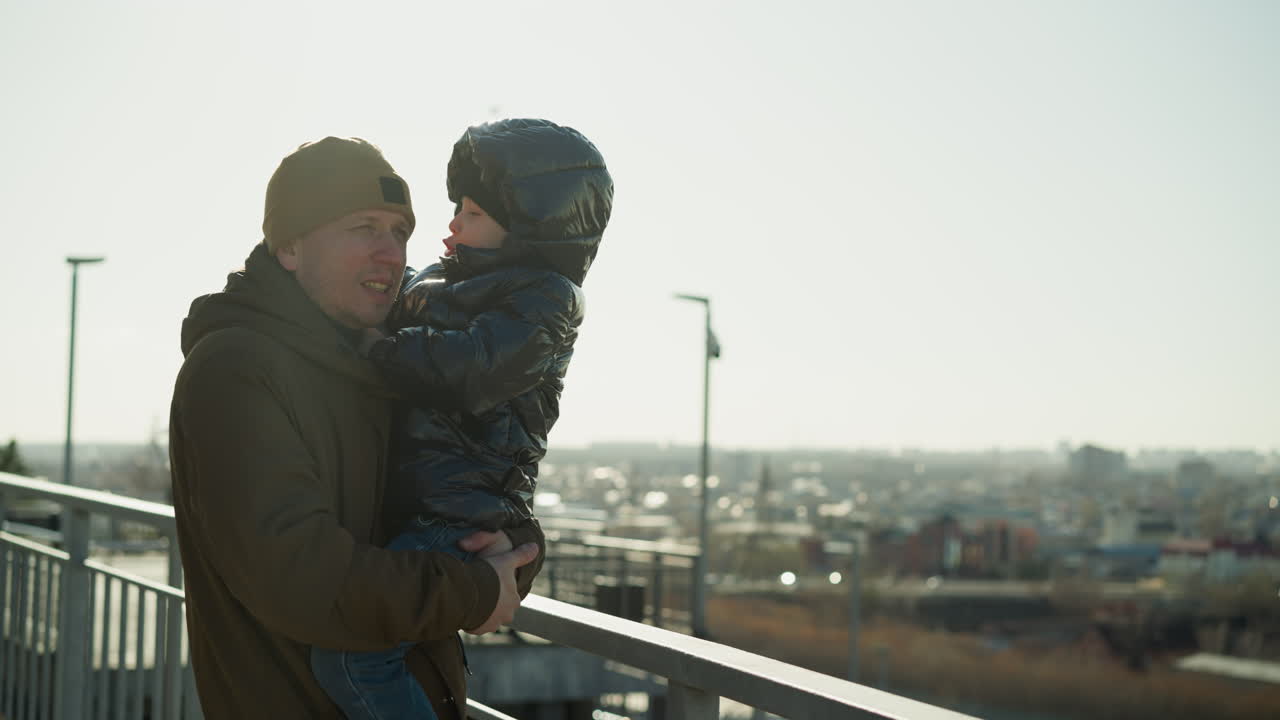 un padre está de pie cerca de una barandilla de metal, llevando a su hijo que suavemente pone su mano en el hombro del padre, mirando hacia un paisaje urbano en la distancia