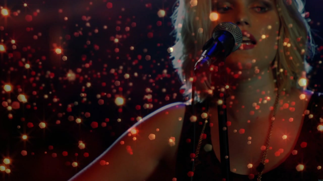 Camera pulling back, performer moving guitar toward mic, singing music with gold overlay particles