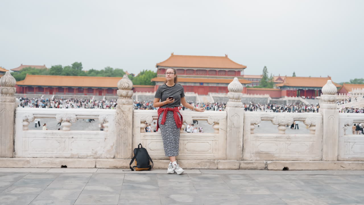 Frau steht an der Palastbalustrade mit Blick auf die Verbotene Stadt, verziertes Steingeländer, entfernte Tempeldächer, belebter Innenhof, Rucksack am Boden, nachdenkliche Pose, bewölkter Himmel, Reisestimmung.
