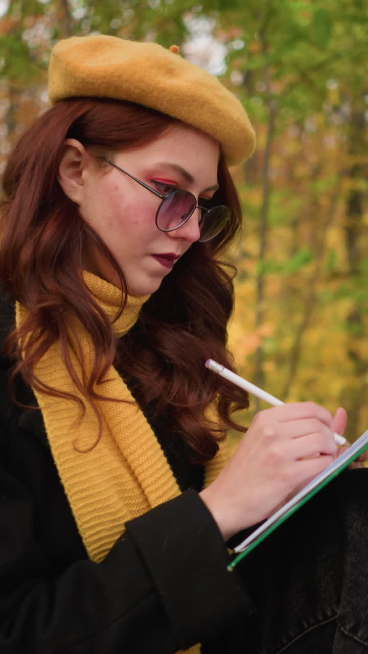 artista con boina amarilla y silenciador profundamente enfocada en su dibujo sentada en un banco rodeada de hojas de otoño, encarnando la concentración artística en un entorno natural