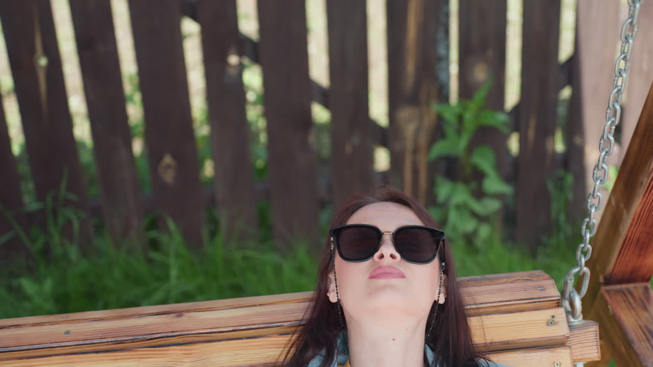 Young girl leans back on wooden swing wearing sunglasses, enjoying quiet outdoor moment, head tilted upward, eyes closed in relaxation, surrounded by lush grass and wooden fence in warm backyard