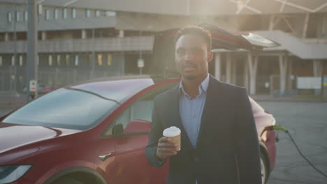 african man drinking coffee while electric car is charging handsome african man in business suit