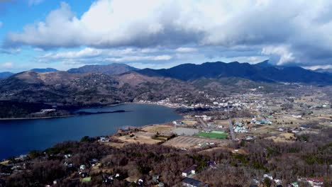 a serene lakeside town surrounded by mountains and clouds, casting shadows on the landscape, aerial view