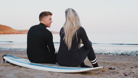 rear view of couple wearing wetsuits sitting on surfboard and looking out to sea together