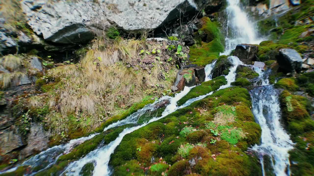 Static drone view over waterfall in Bucegi National Park, Romania, focusing on water splitting into multiple streams as it flows over moss-covered rocks, with blue water and gray-white stones