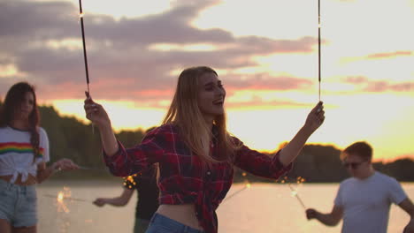 la mujer rubia con camisa a cuadros roja y pantalones cortos de vaqueros está bailando con grandes luces bengalíes en sus manos en la playa con sus amigos.