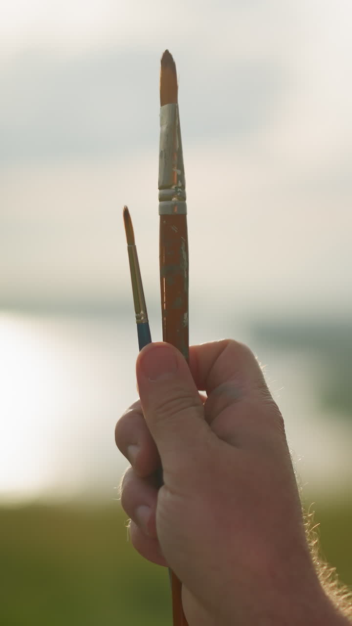 un primer plano de una mano sosteniendo dos pinceles uno pequeño y otro muy pequeño contra un fondo soleado y borroso. la iluminación suave enfatiza las herramientas del artista, capturando un momento de creatividad
