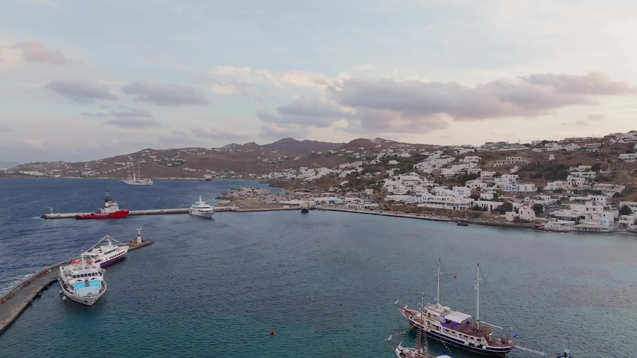 Ripresa panoramica con drone del vecchio porto di Mykonos all'alba, Cicladi, Grecia. Barche ormeggiate nel porto, nuvole sparse, luce dorata del mattino