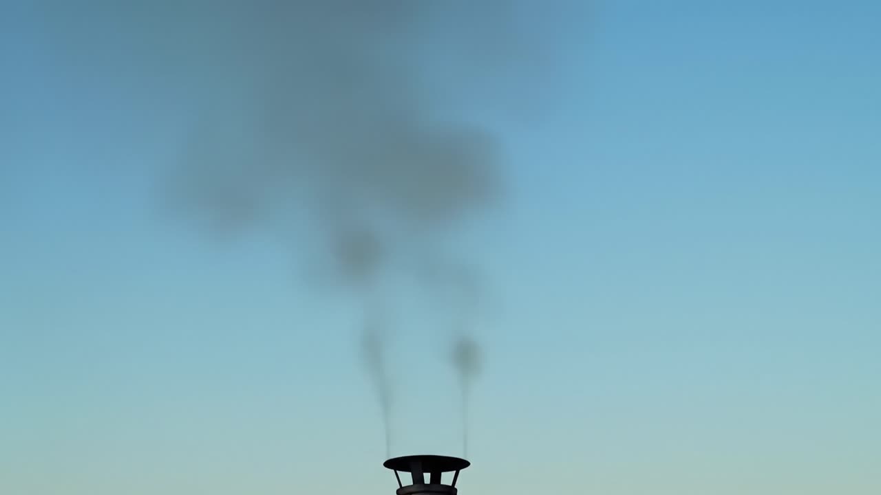 Smoke billows from a chimney as the sun sets in the background, creating a striking contrast against the clear blue sky. The scene evokes a peaceful evening ambiance.