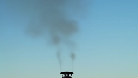 smoke billows from a chimney as the sun sets in the background, creating a striking contrast against the clear blue sky. the scene evokes a peaceful evening ambiance.