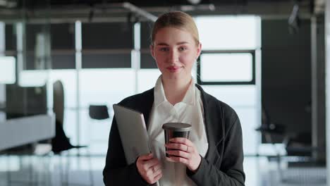 Portrait-of-a-happy-blonde-girl-in-a-business-suit-who-holds-a-cup-of-coffee-and-a-laptop-in-her-hands-while-working-in-a-modern-office