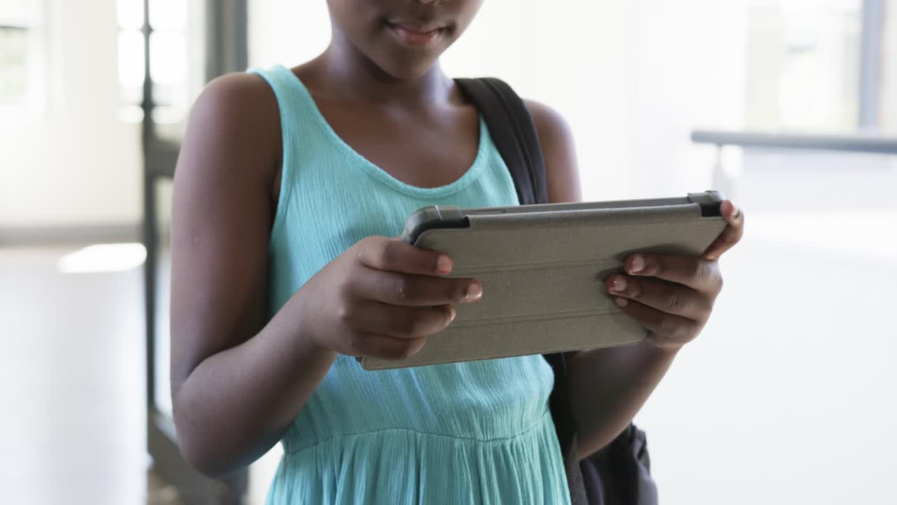 African American child standing in bright hall with bag holding tablet, tapping to do homework