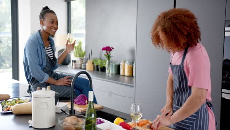 happy diverse couple wearing aprons drinking wine and preparing meal in kitchen, slow motion