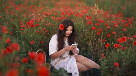 close-up-beautiful-dark-haired-girl-with-a-poppy-in-her-hair-sitting-in-a-field-of-wildflowers-and-red-poppies,-wearing-a-dress,-smiling-while-playing-a-game-on-her-smartphone