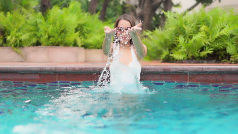 Asian-woman-in-white-bathing-suit-and-large-sunglasses-splashes-water-into-the-air-with-her-hands-while-standing-in-pool-with-tropical-plants-in-background