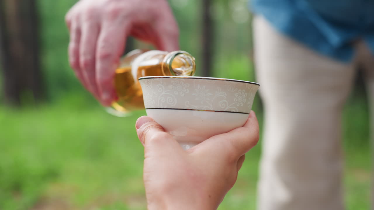 화창한 녹색 공원에서 편안한 야외 피크닉을 즐기는 동안 어린 소녀가 들고 있는 흰색 컵에 남성의 손이 상쾌한 레모네이드를 따르는 모습을 가까이에서 보여주며, 따뜻한 가족 간의 연결과 평화로운 여름날의 한때를 나타내요.