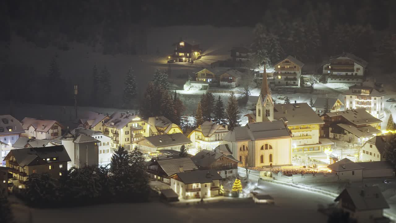 The church spire rises from a warmly lit Tyrolean village on a crisp winter night. Tilt-shift video.