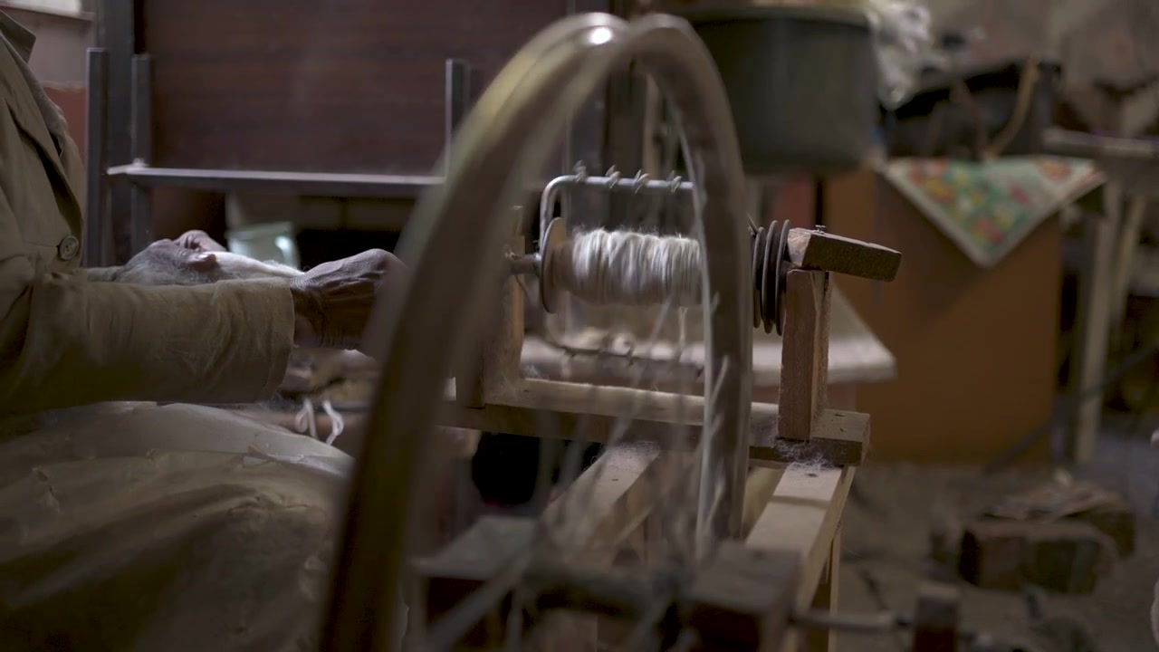 An African woman artisan's hands expertly turn Angora wool into yarn on a spinning wheel in a dimly lit workshop. Ideal for cultural heritage and craft-focused content.